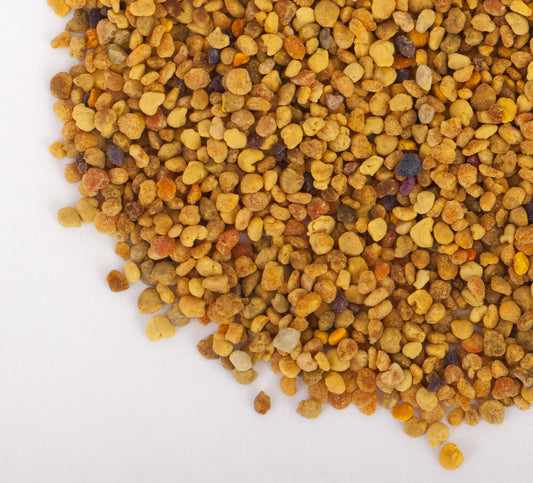 A close-up image of a 2.5 oz pile of ochre-colored Honeybee Bee Pollen scattered over a white surface. The bee-collected granules vary in size and color, ranging from yellow and orange to light brown and purple.