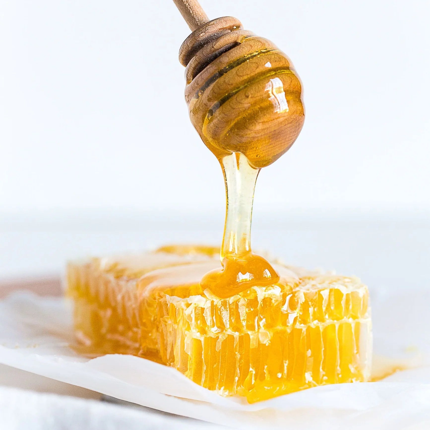 A wooden honey dipper drizzles golden honey over a block of 100% edible Honeybee Honeycomb 1 lb. set on white parchment paper. The honey glistens as it cascades down, showcasing the rich texture and depth of the pure raw honeycomb—perfect for charcuterie boards.