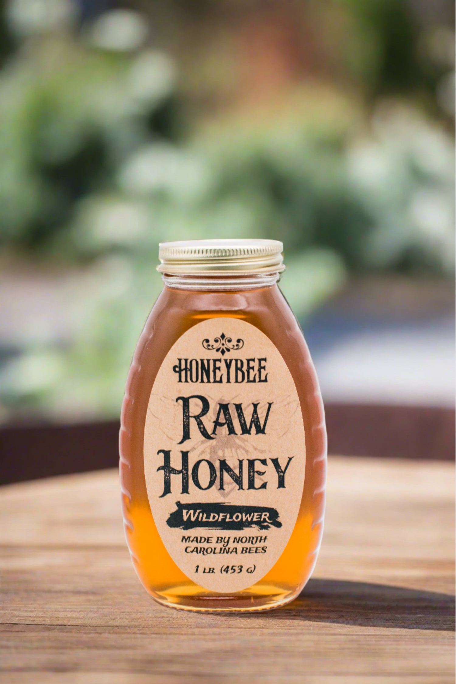 A glass jar containing "Honeybee Raw Honey," labeled as premium honey made by Honeybee, sits on a wooden surface. The jar holds 1 lb (453 g) of all-natural raw honey, with a softly blurred greenery background.