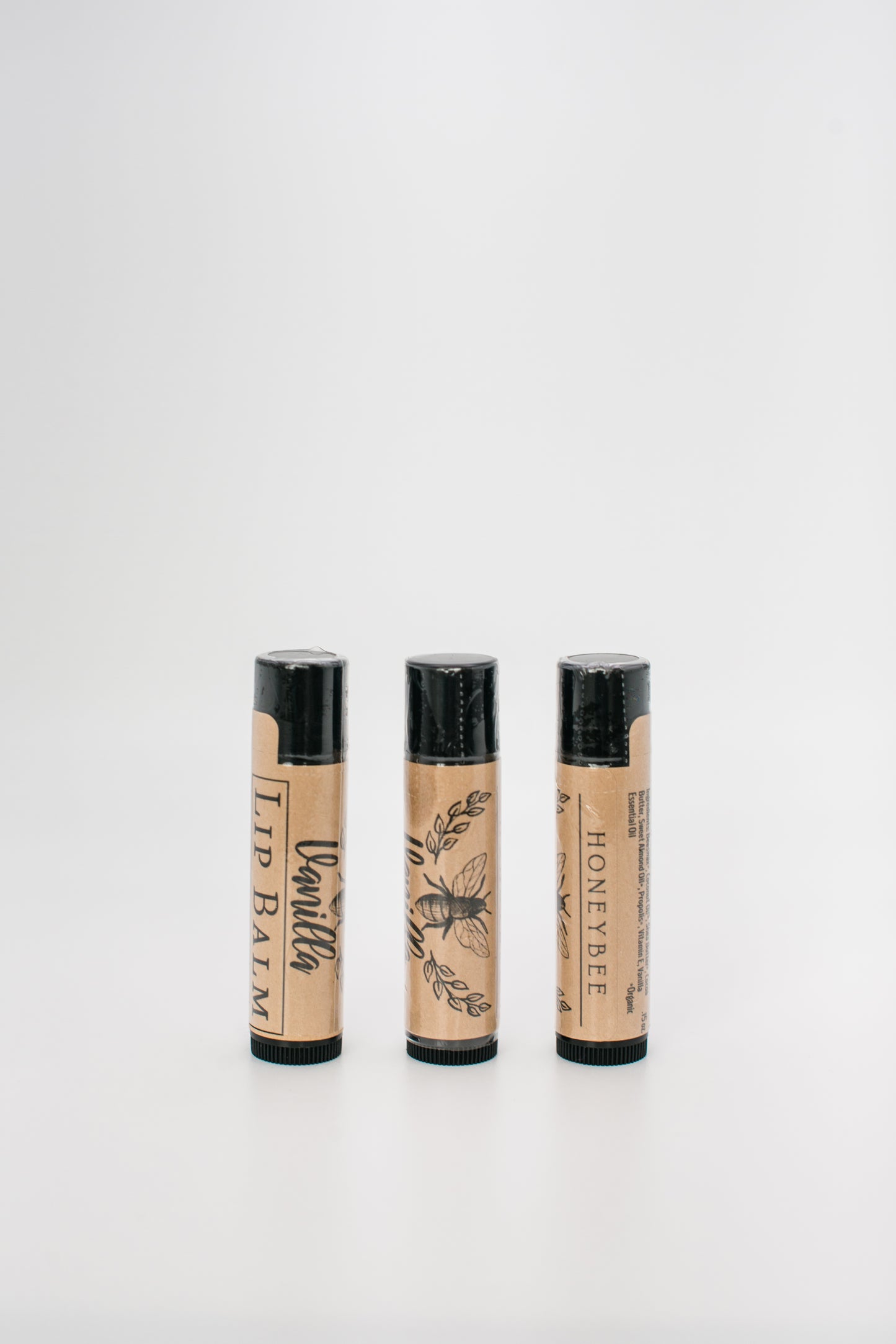 Three tubes of handcrafted lip balm stand upright against a white background. Each tube has a brown, nature-inspired design featuring black text and illustrations. The labels read "Beeswax Lip Balm" and "Honeybee," highlighting their all-natural, beeswax ingredients.