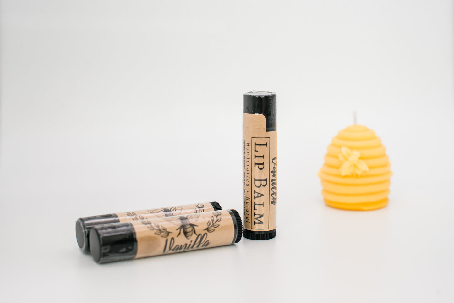 The image shows four tubes of Honeybee Beeswax Lip Balm, vanilla-scented and all-natural, with a brown label featuring a bee logo and black caps. One tube is standing upright, while three lay down. Next to the handcrafted lip balms is a yellow, beehive-shaped candle with a small bee decoration.