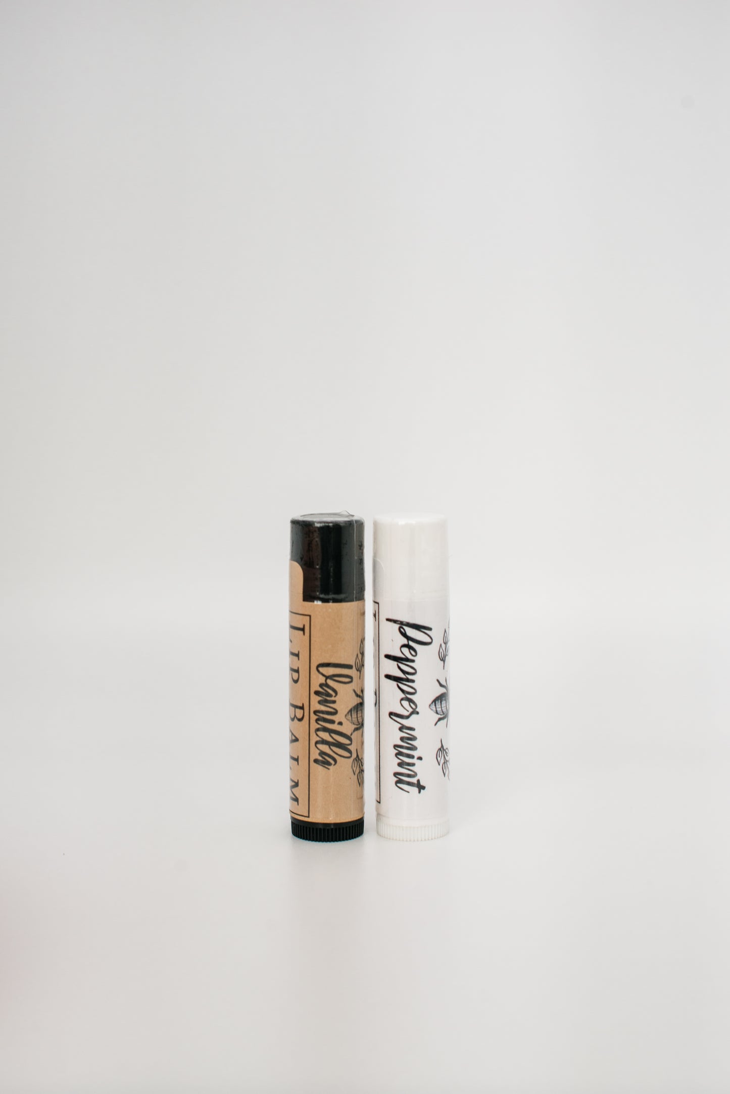 Two handcrafted Beeswax Lip Balm tubes from Honeybee stand upright on a white surface against a plain white background. The left tube, an all-natural vanilla lip balm, has a brown label with black text, while the right tube, labeled as peppermint beeswax lip balm, features a white label with black text.