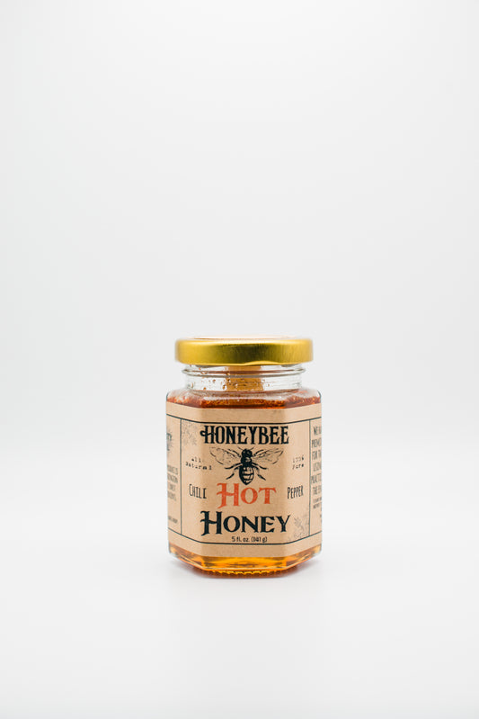 A jar of Honeybee's 'Hot Honey' with a gold lid set against a plain white background. The label features an illustration of a bee and emphasizes that the raw wildflower honey is infused with hot chili peppers, suggesting a spicy flavor.