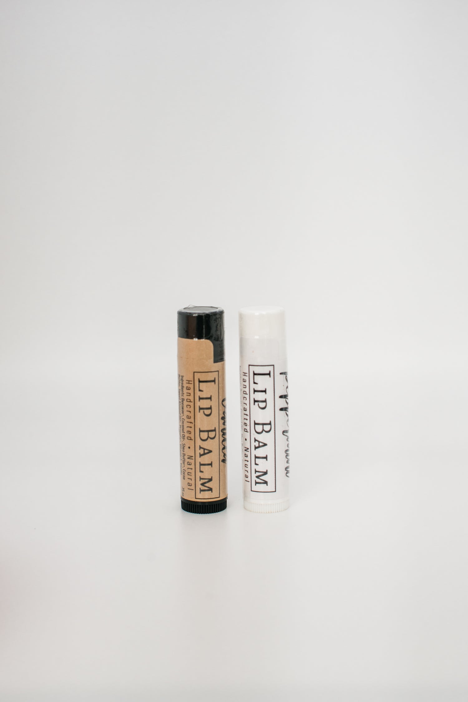 Image of two lip balm tubes standing upright against a plain white background. The handcrafted tube on the left has a black cap and tan label, while the Beeswax Lip Balm from Honeybee on the right has a white cap and white label. Both all-natural tubes are labeled "LIP BALM.