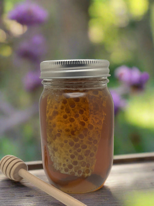 A glass jar filled with Honeybee Chunk Honey sits on a wooden table. A wooden honey dipper is placed beside the jar. The background is softly blurred, showing purple flowers and greenery, hinting at a garden setting.